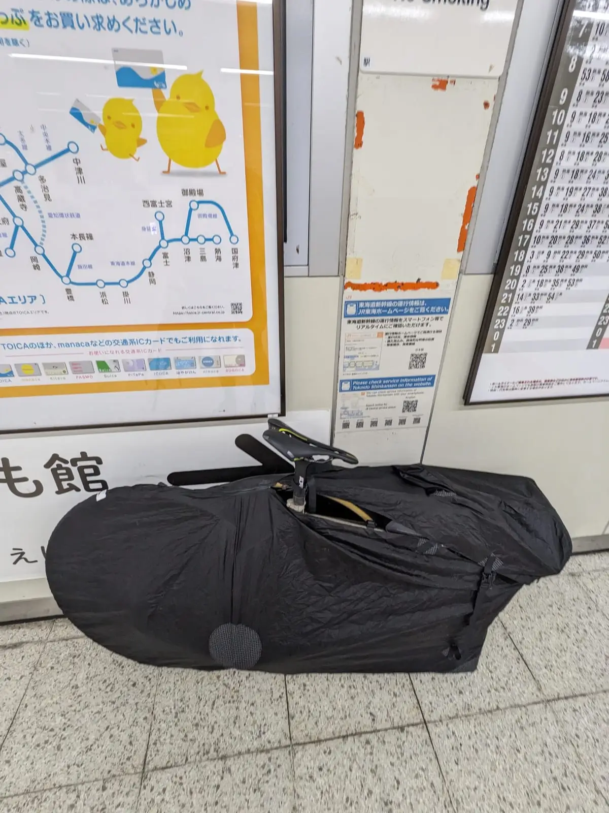 The bike packed into its rinko bag, waiting on a Japanese train platform beside route maps