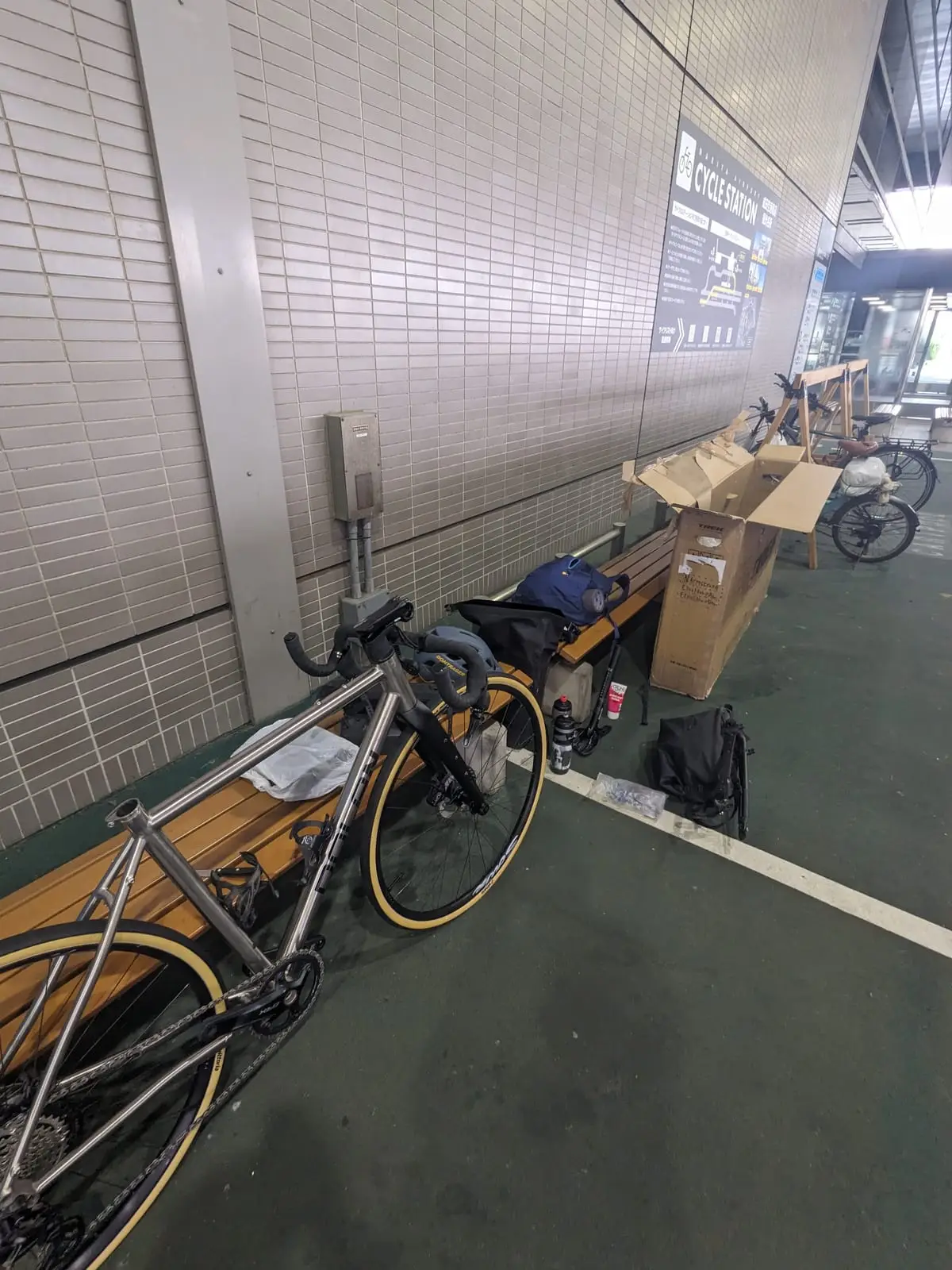 A bicycle frame propped against a bench mid-rebuild in an airport corridor, with a cardboard bike box and tools nearby