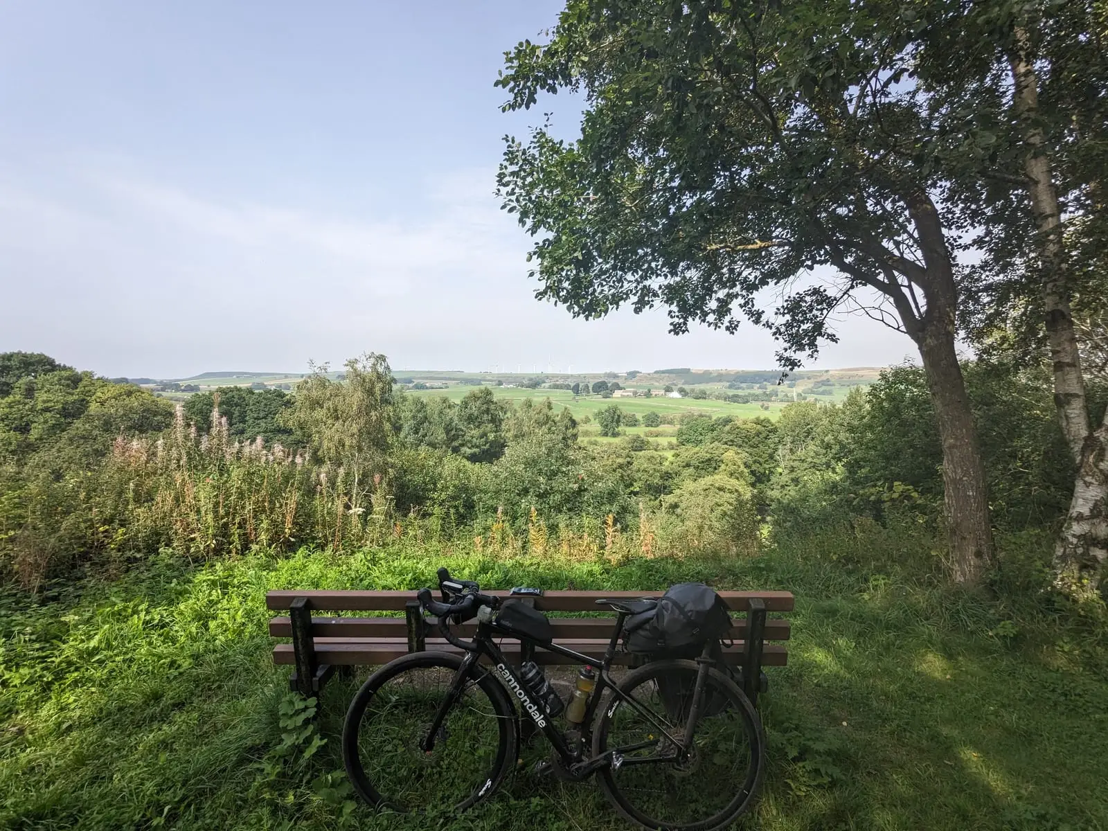 A touring bike leaning against a wooden bench under a tree, looking out over a green valley