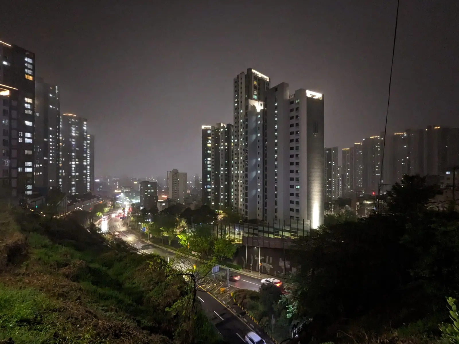 A row of illuminated Busan apartment towers at night, seen from a hillside above a busy road