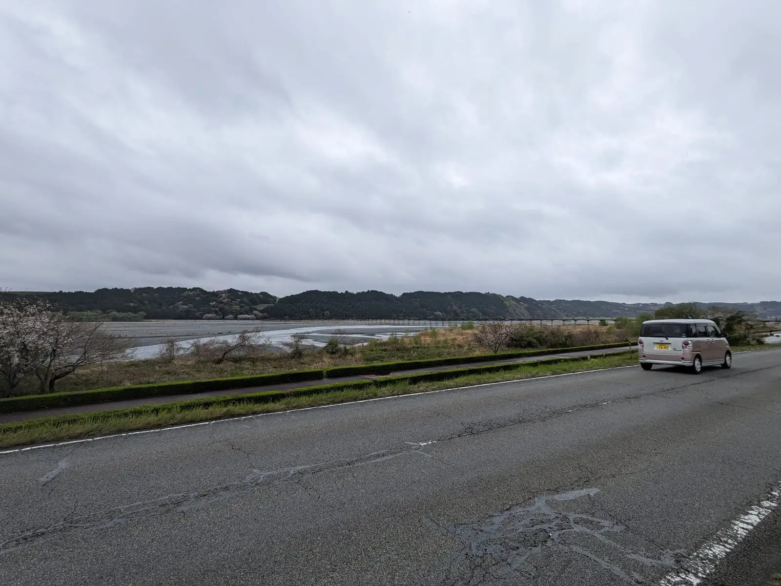 A wide, calm estuary beside a straight coastal road under an overcast sky, with a van parked at the roadside