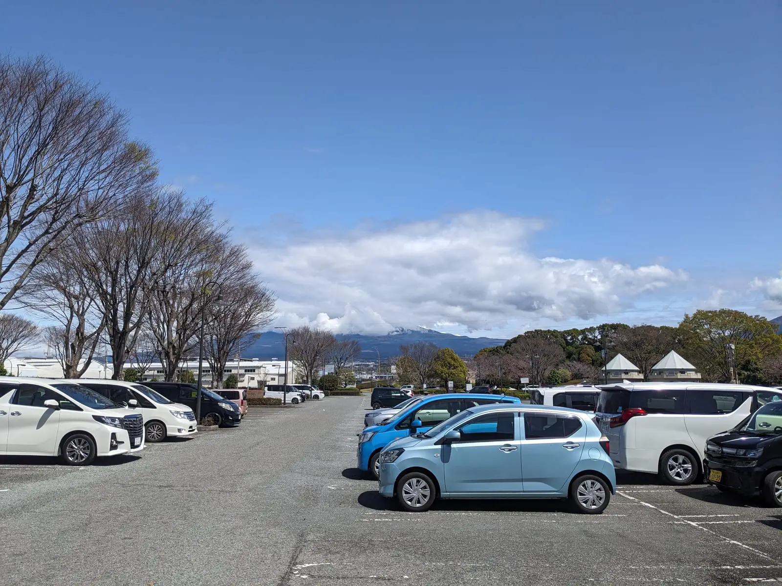 A café car park with several cars parked and hills visible in the distance, Mount Fuji still hidden by cloud
