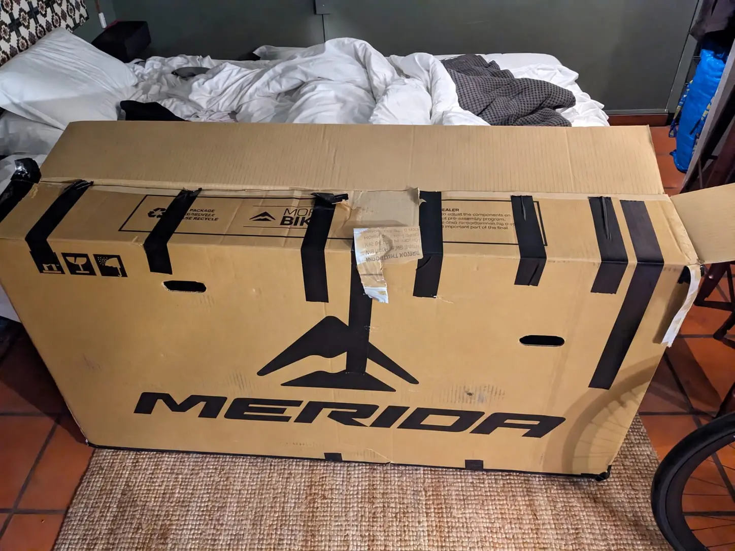 A large cardboard bike box in front of an unmade bed in a Porto room