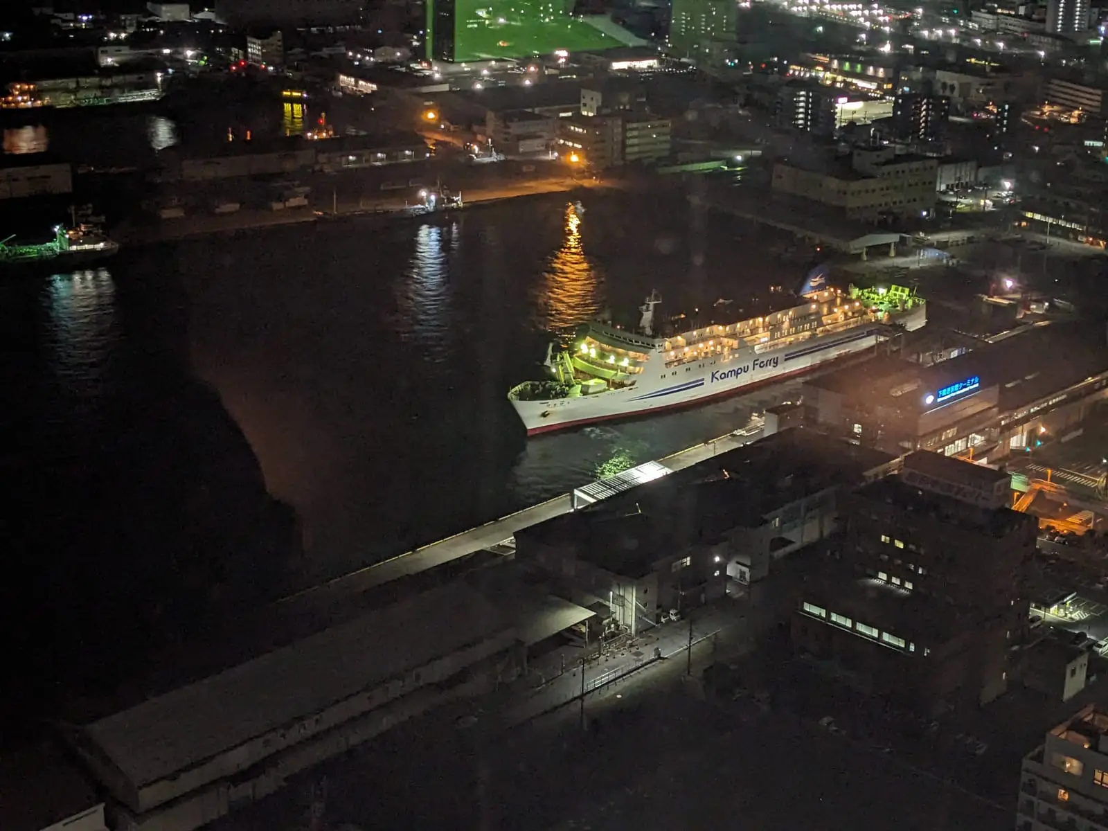 A night view over a harbour with a brightly lit ferry on dark water, seen from a height