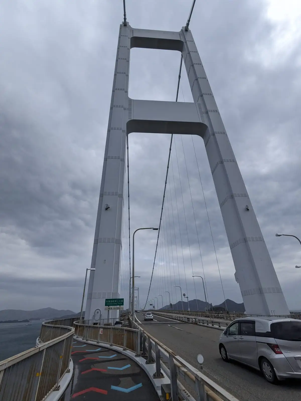 A tall white suspension tower of the Kurushima-Kaikyo Bridge against a cloudy sky, with the cycle path curving below and a car crossing the road deck