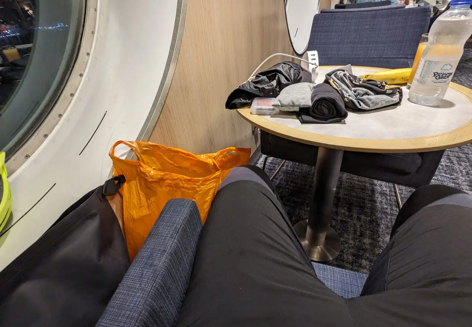 A pair of legs stretched across two ferry seats, with an orange bag and a table of cycling kit beyond