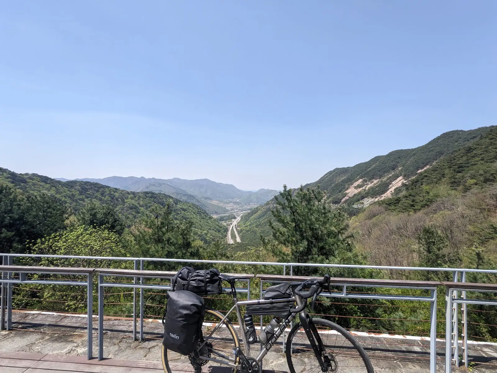 A loaded touring bike resting against a railing at a viewpoint over a layered green mountain valley, with a road winding through it below