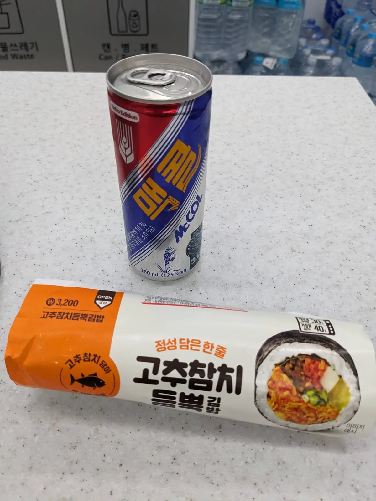 A can of Maccol soft drink beside a spicy rice-cake snack on a convenience-store shelf