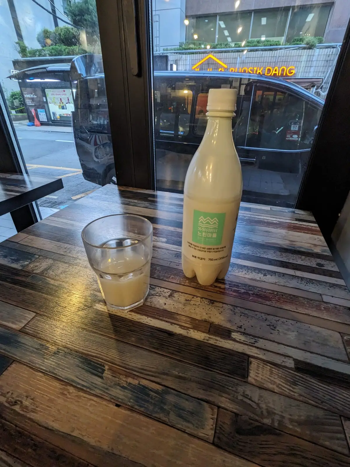 A bottle and glass of cloudy makgeolli rice wine on a wooden café table by a window