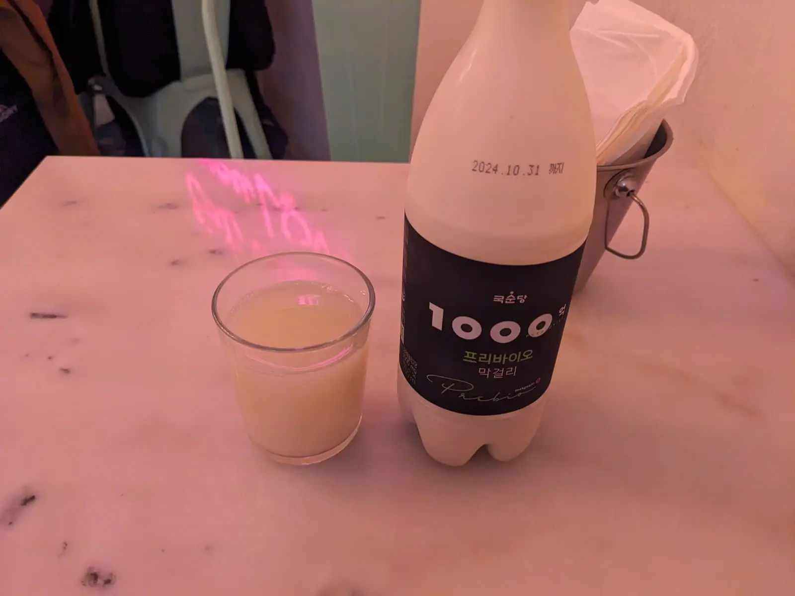A bottle and glass of makgeolli on a marble table under pink light