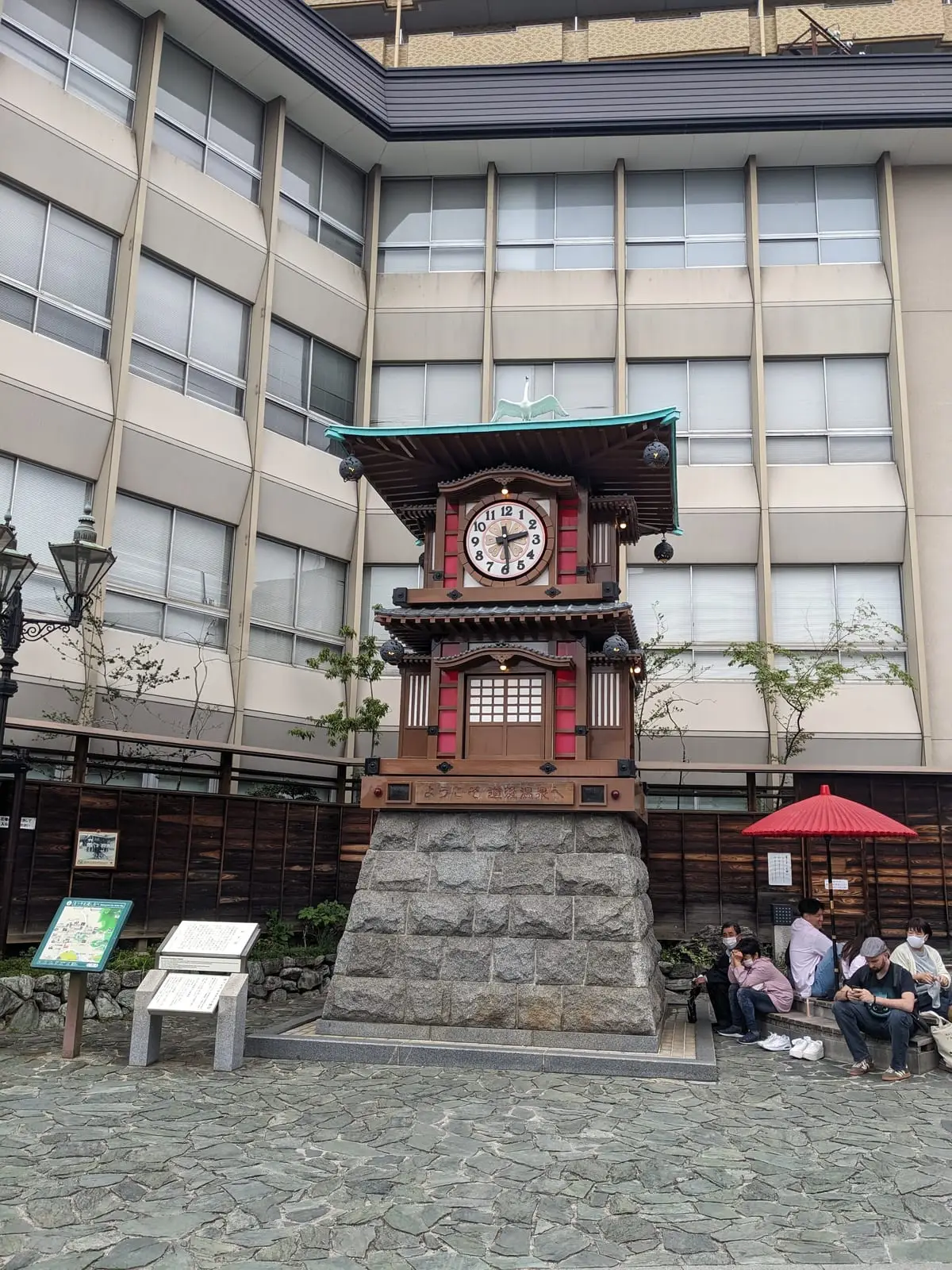 An ornate wooden karakuri clock tower on a Matsuyama street corner, with a red umbrella and modern buildings behind