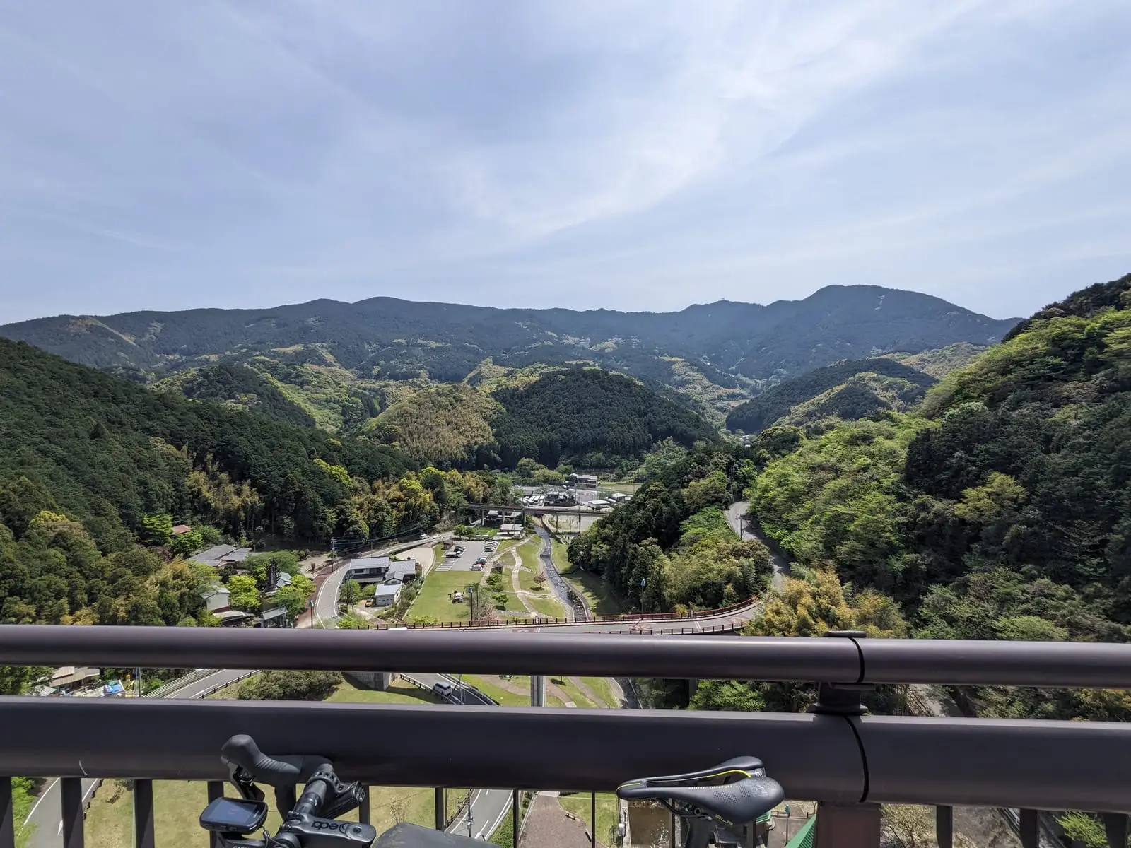 A mountain valley and village seen from a road along the top of a dam, with a guardrail in the foreground