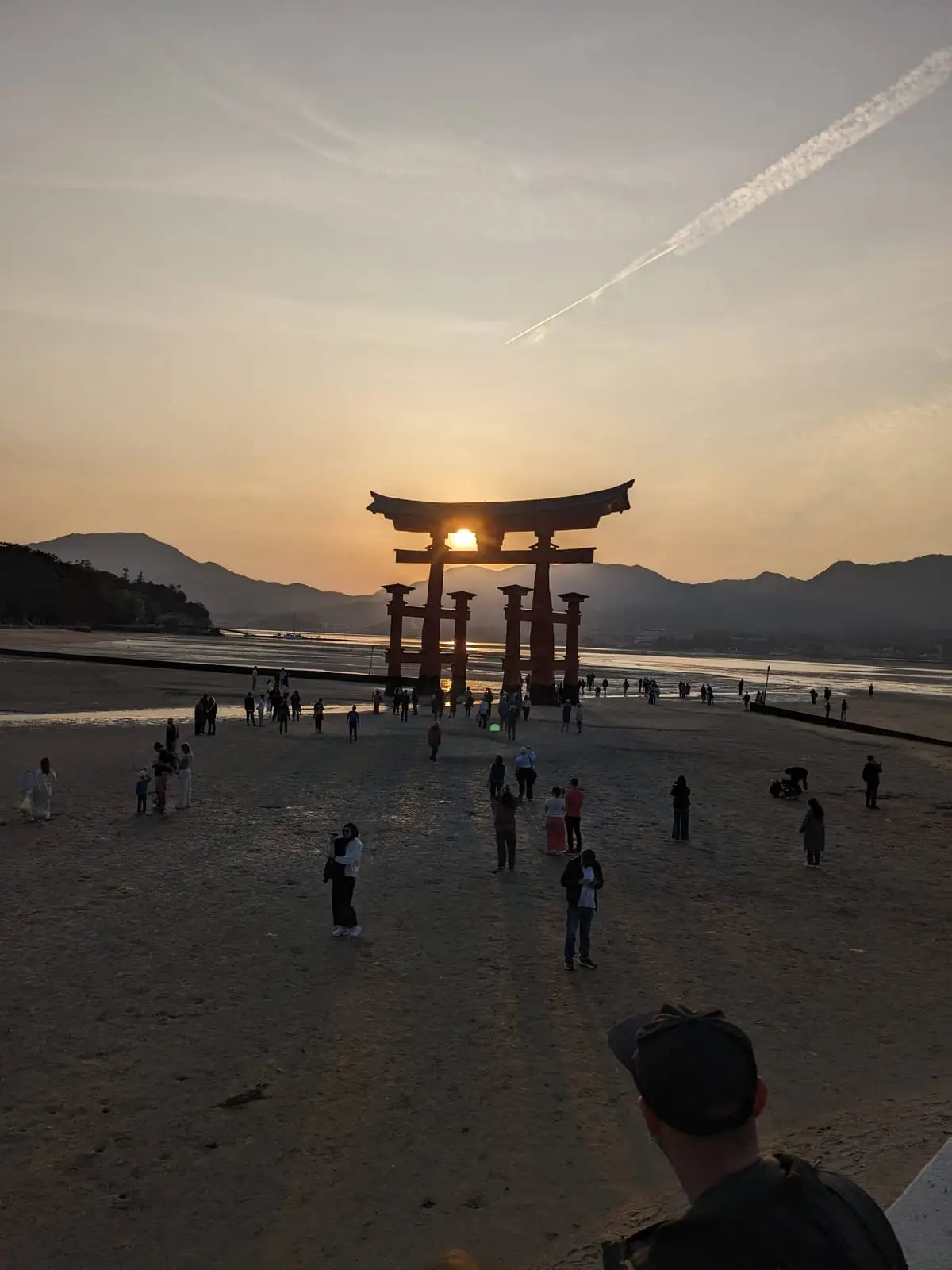 The Otorii gate silhouetted against a vivid orange sunset, with visitors gathered on the sand nearby
