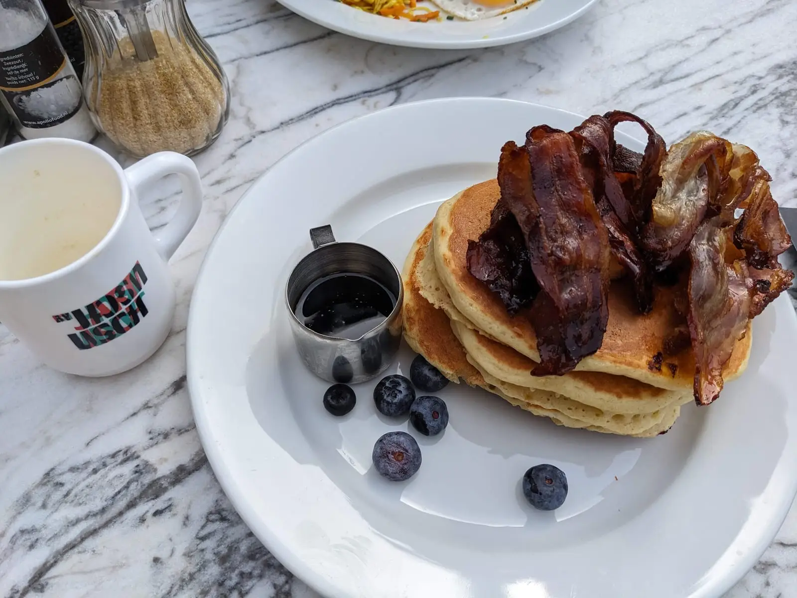 A stack of pancakes topped with crisp bacon, blueberries, and a small jug of syrup