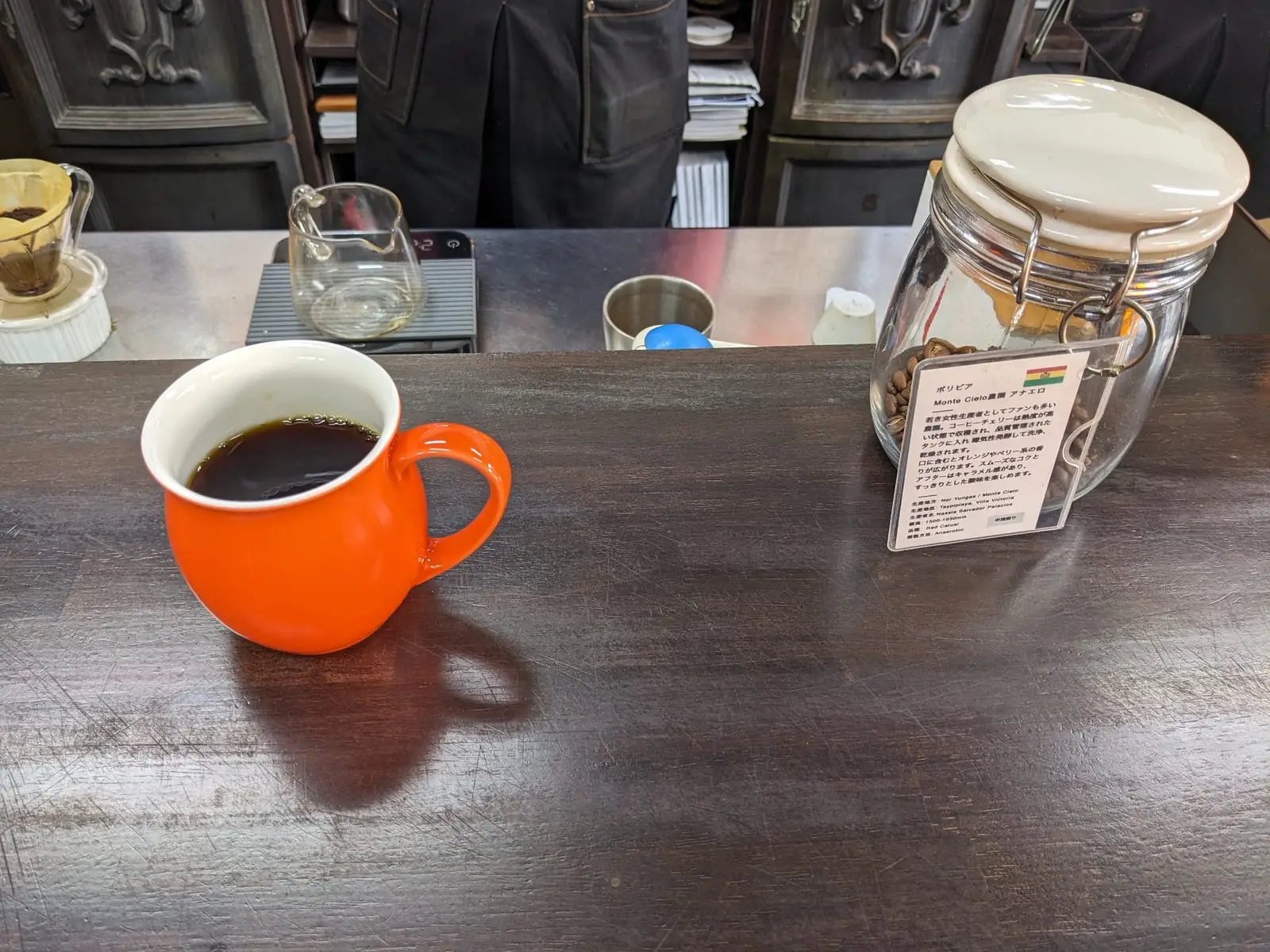 A pour-over coffee setup with glass carafes and an orange mug of coffee on a wooden counter at Ichi Coffee Roastery
