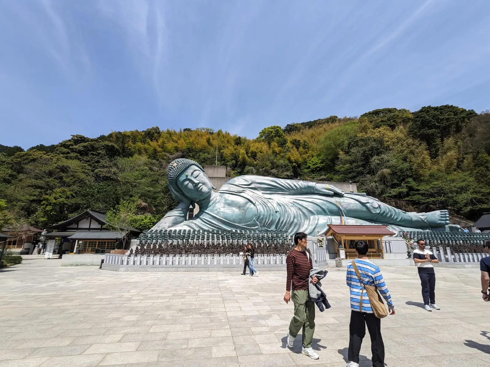 A large reclining Buddha statue on a paved plaza, with several visitors standing in front and a wooded hillside behind