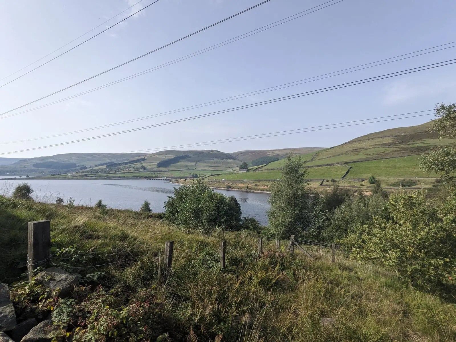 A reservoir in a green Peak District valley beneath power lines and a pale sky