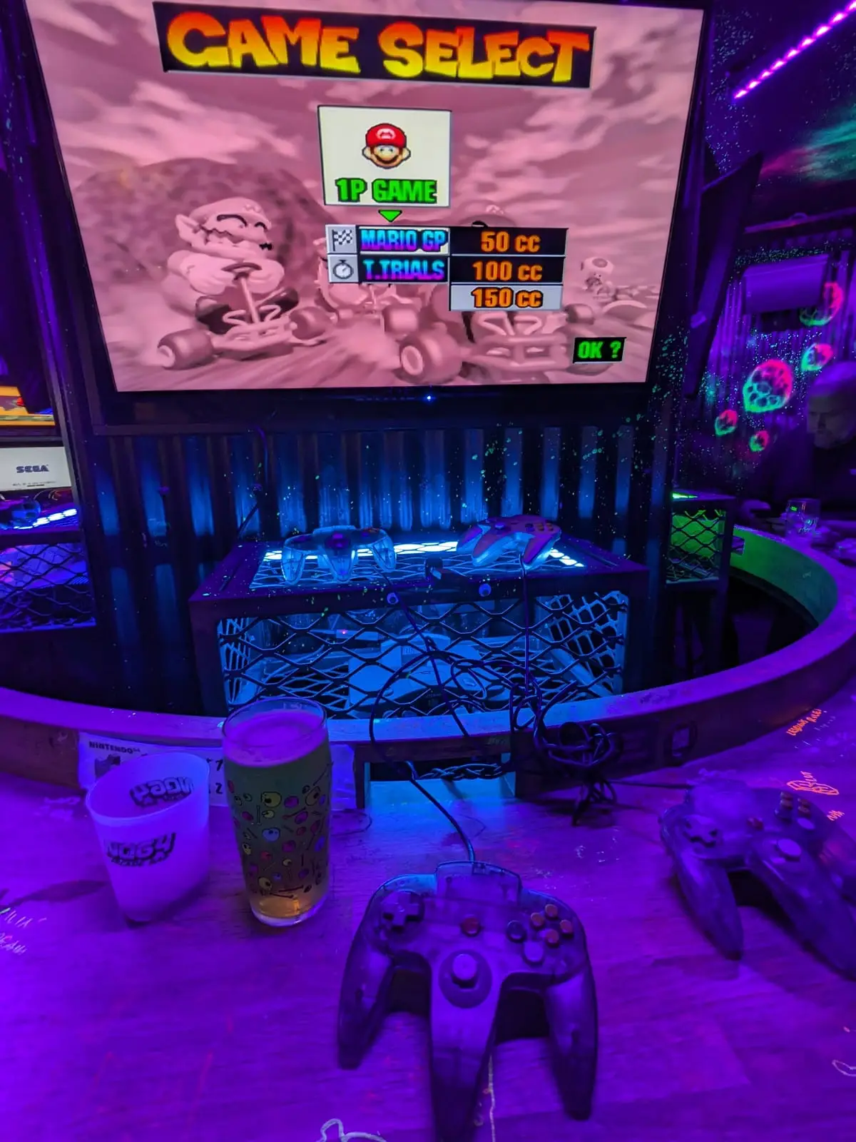 A retro fighting game running in a neon-lit video game bar, with a controller and drinks on the table