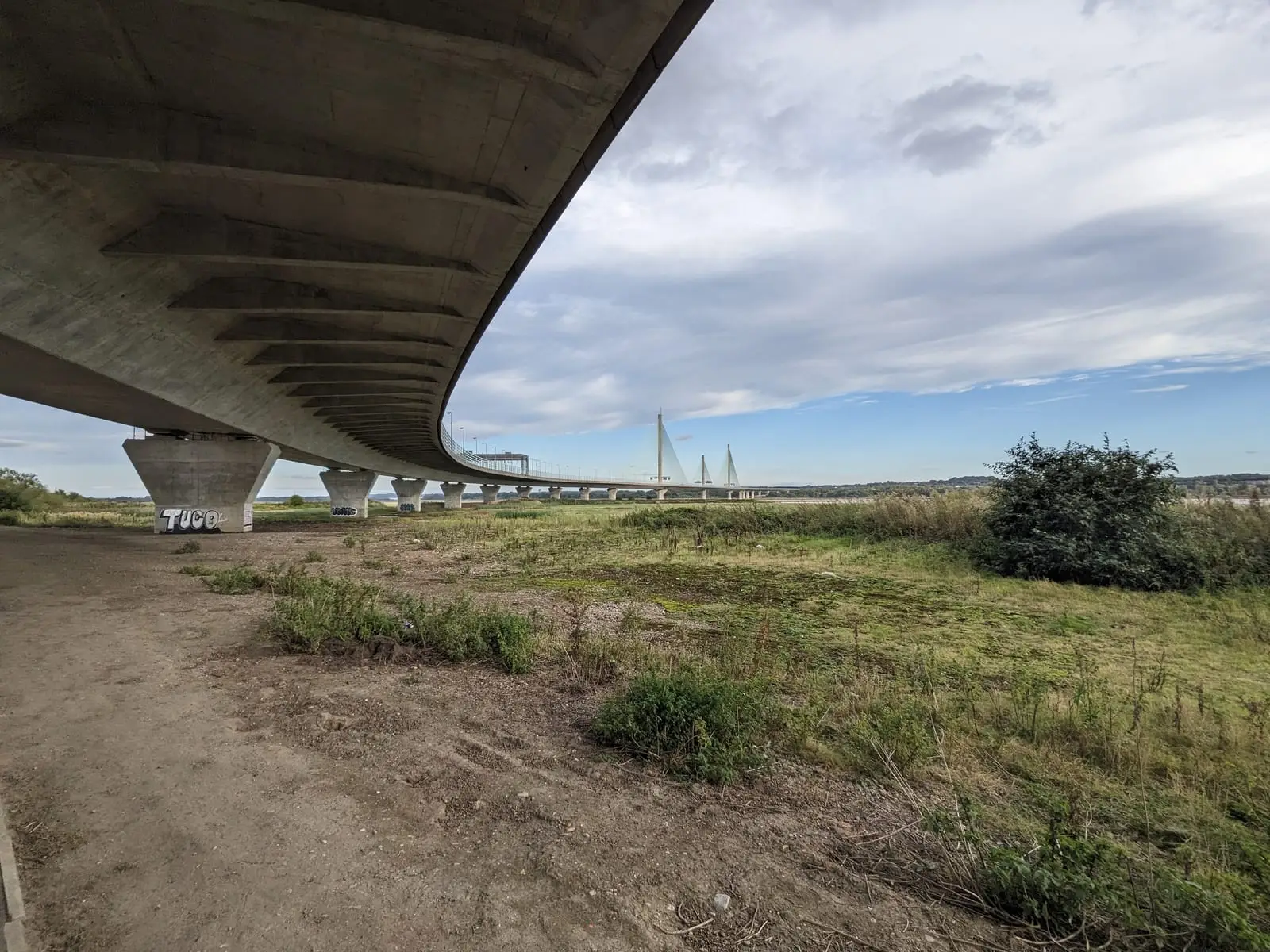 The underside of a large road bridge crossing open ground beside the Mersey