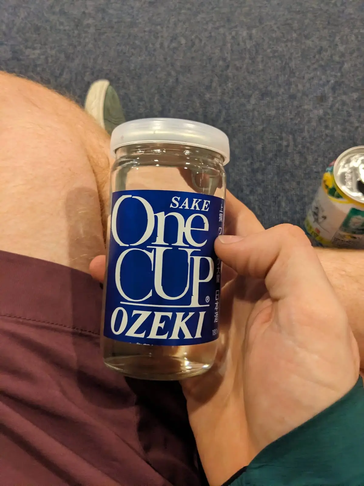 A hand holding a small cup of One Cup Ozeki sake, with other canned drinks blurred on a table behind
