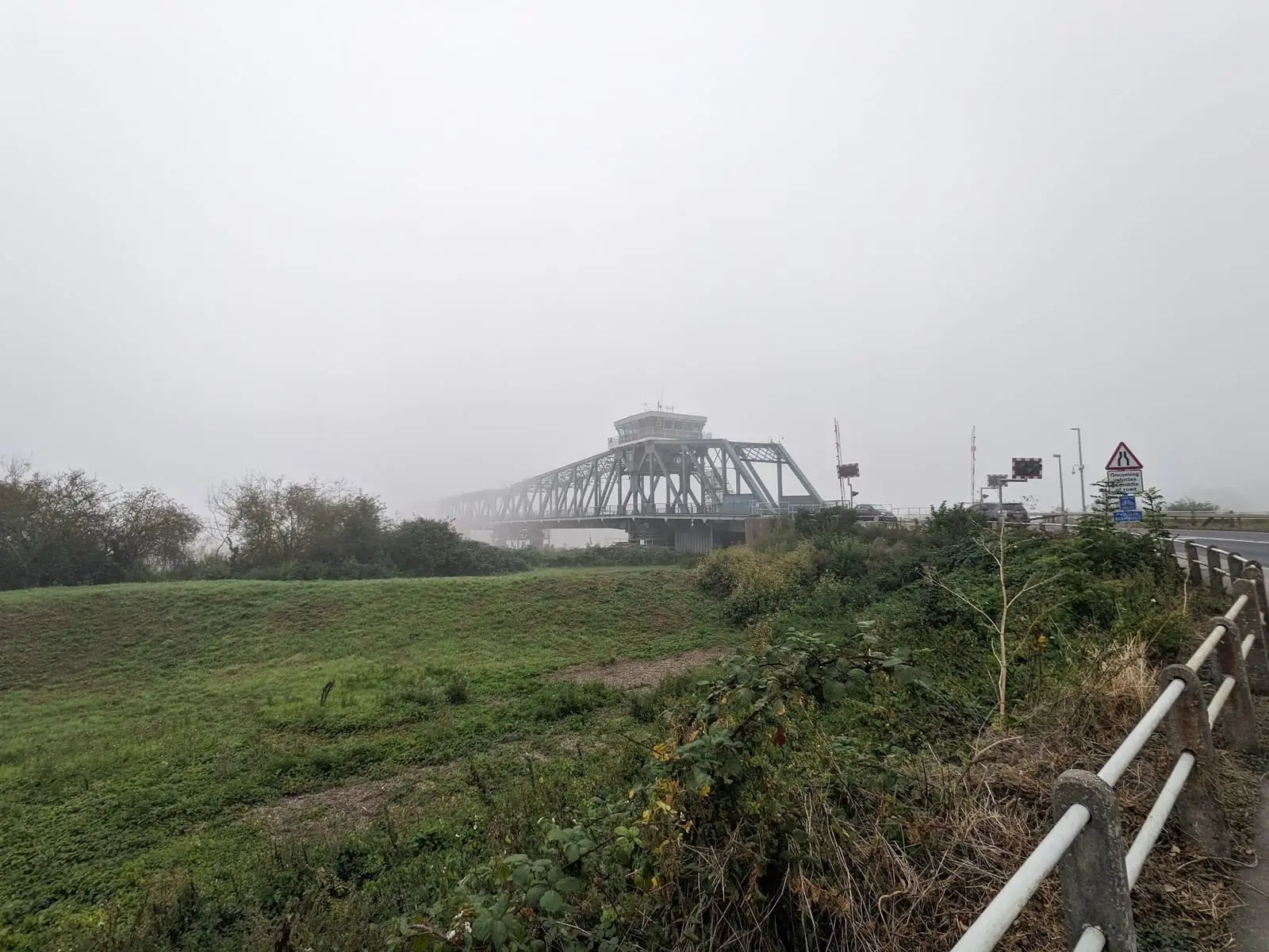 A steel bridge fading into thick fog beyond a grassy bank