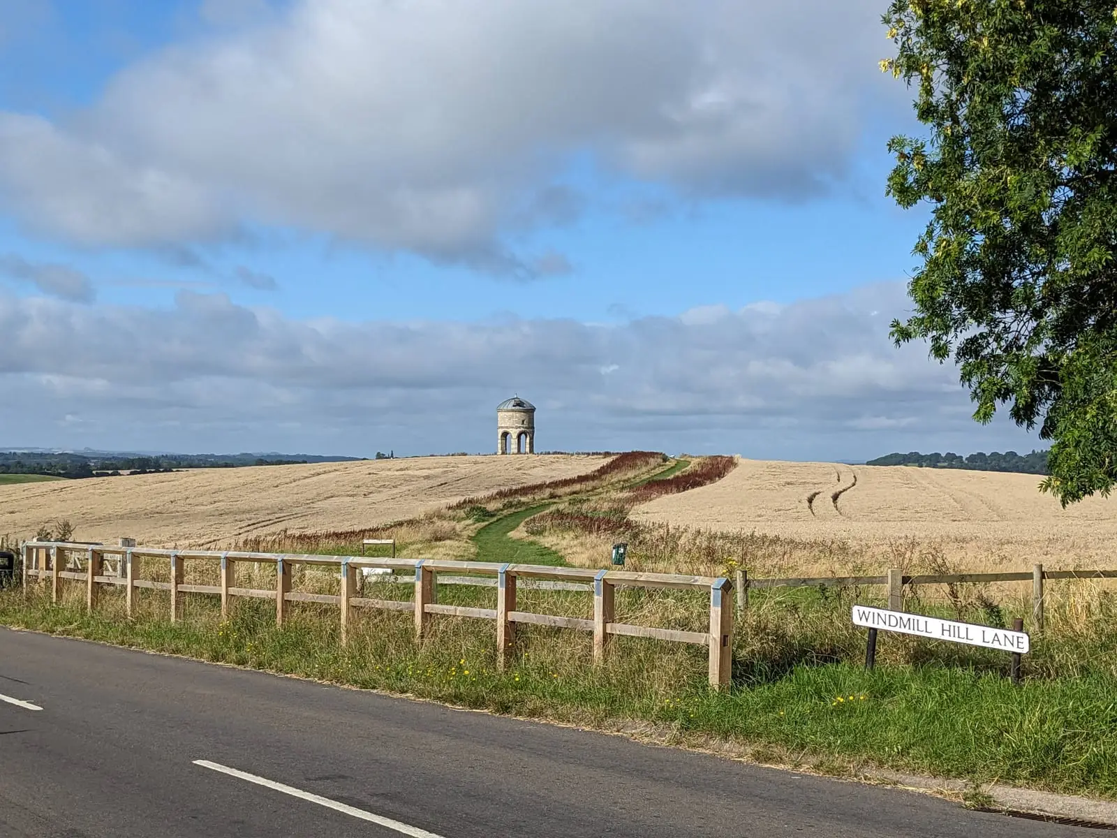 A round stone tower on a hilltop above golden wheat fields, seen across a quiet country road