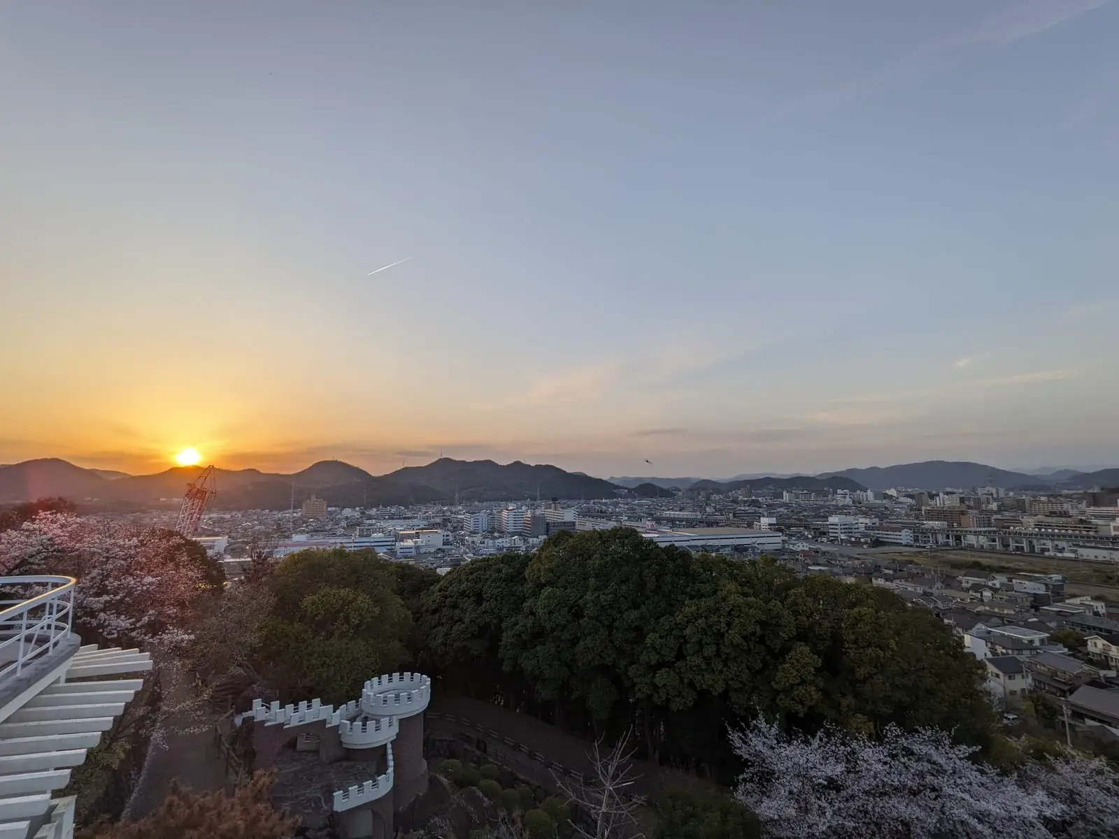 A sunset over a hillside town, with silhouetted buildings and trees in the foreground and the sun low on the horizon