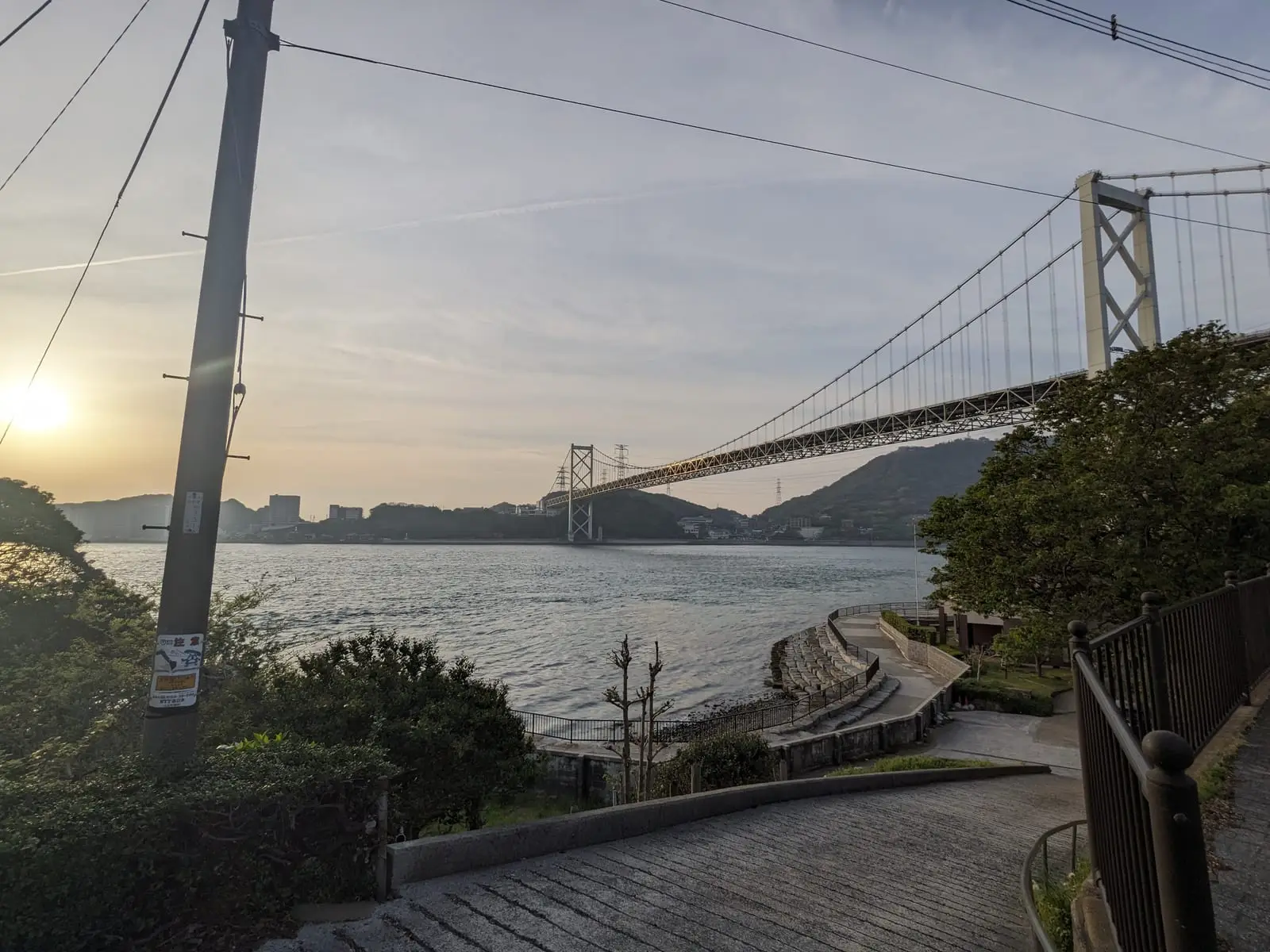 A suspension bridge crossing water at dusk, seen from a hillside path curving down towards it