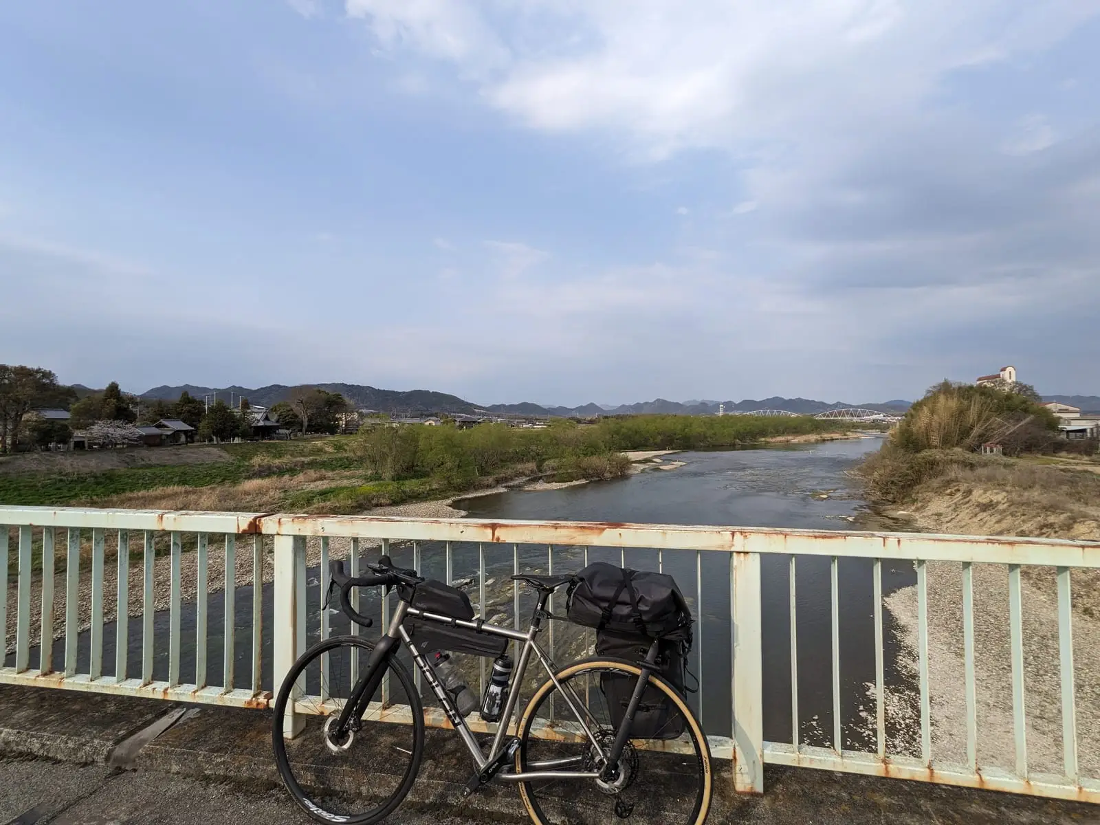 A loaded touring bike propped against a railing beside a river, with hills in the background