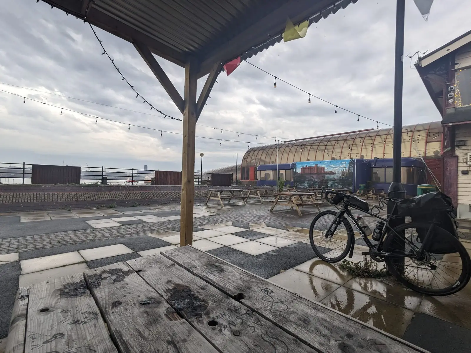 A loaded touring bike on a wet paved terrace under string lights, with an overcast river behind