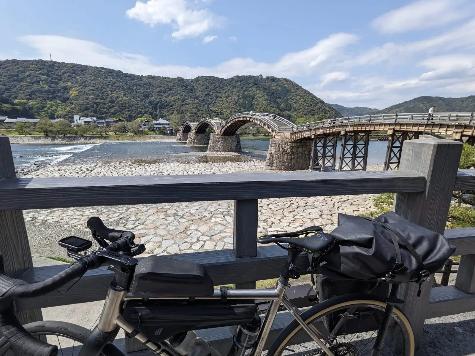 A touring bike leaning against the railing overlooking an arched wooden bridge crossing a wide river.