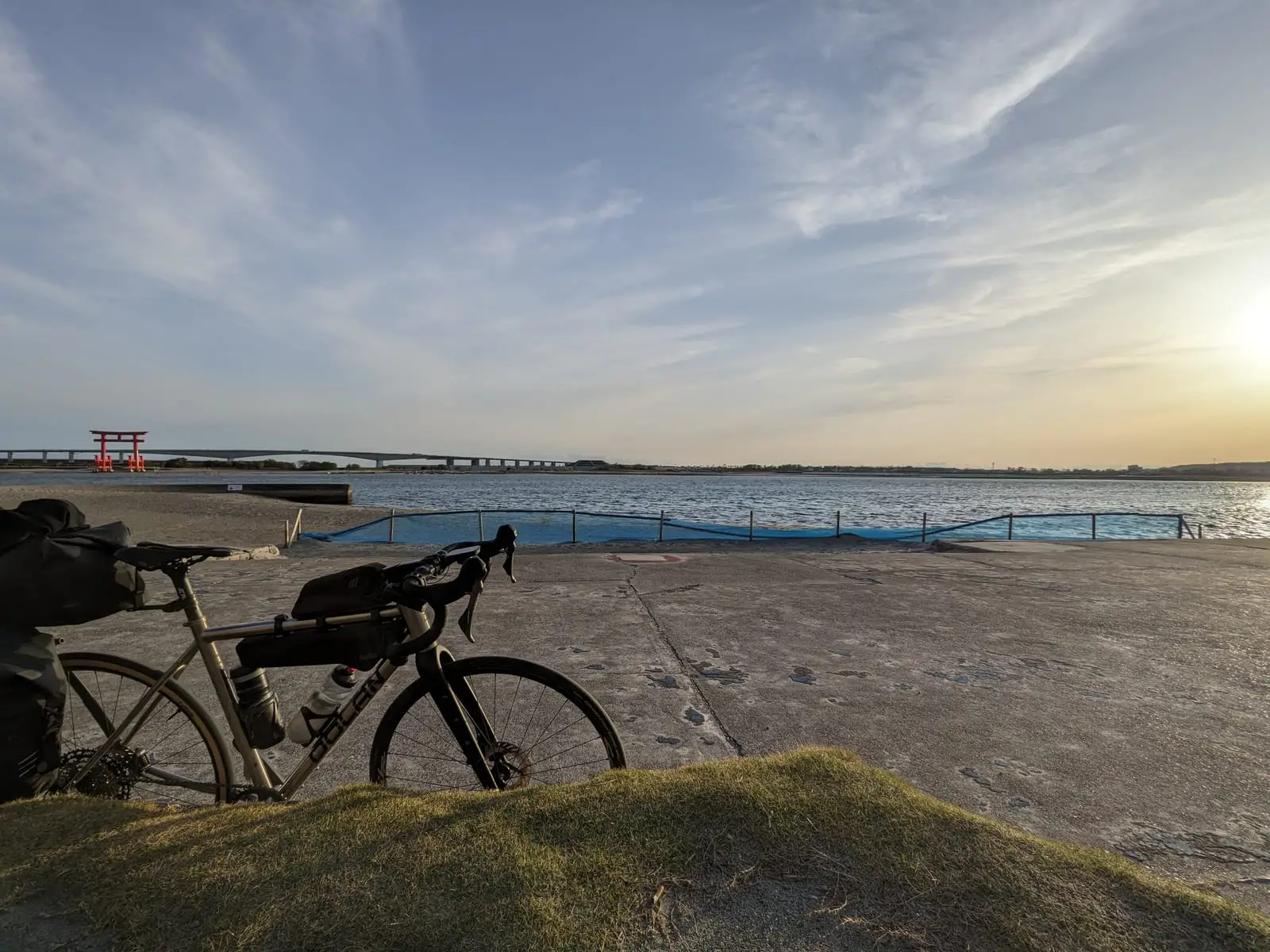 A loaded touring bike parked on a grassy bank overlooking a wide bay, with a red torii gate standing in the water