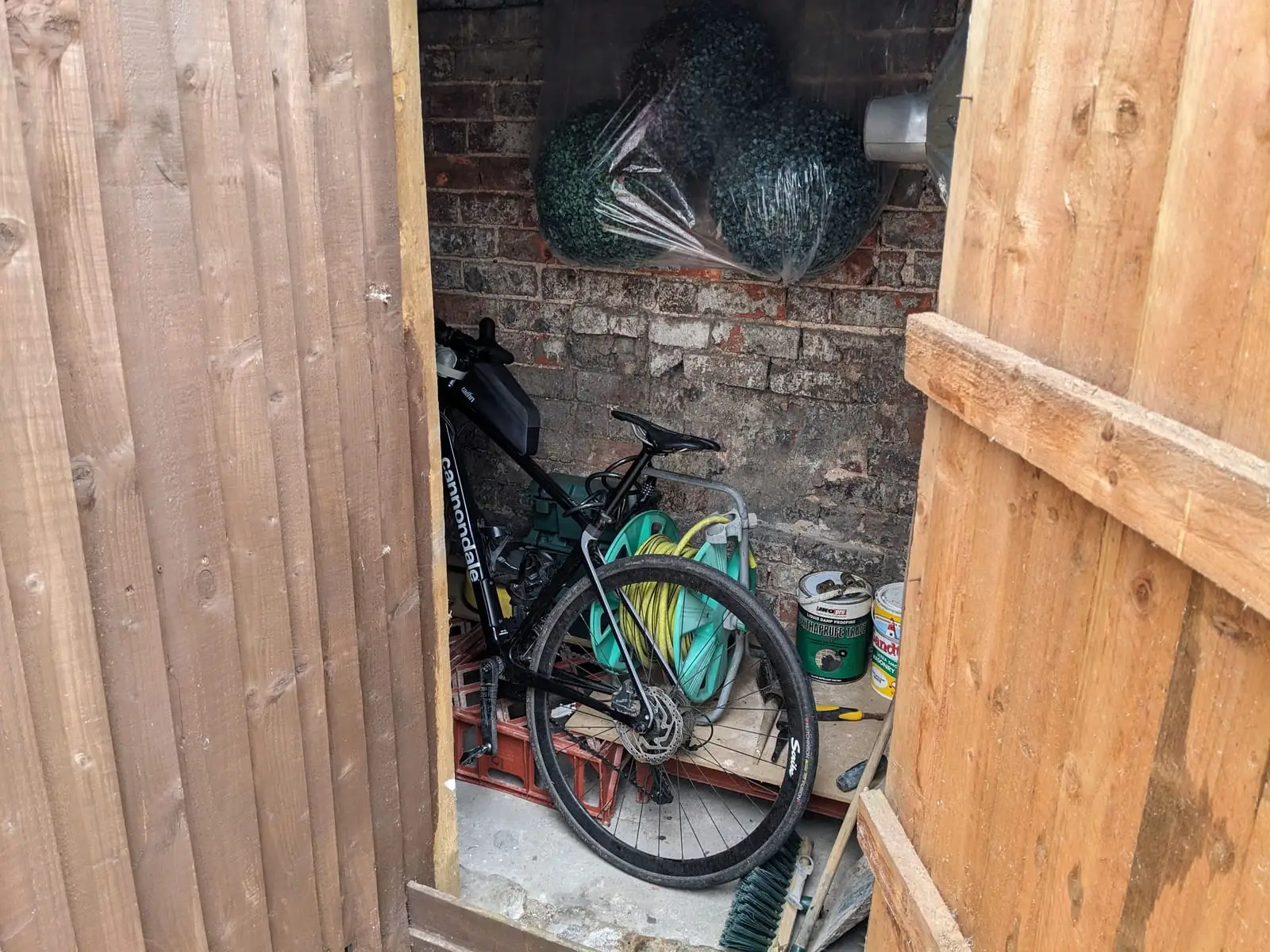 A touring bike propped inside a small brick-walled garden shed among tools and a hose reel