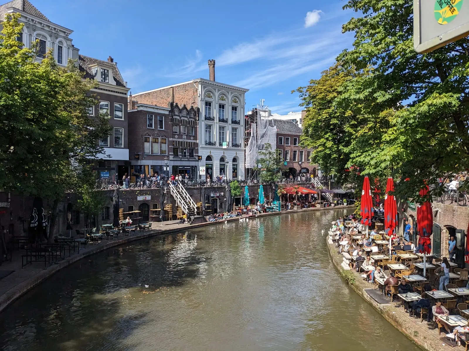A sunlit Utrecht canal lined with waterside cafes and red parasols under a blue sky