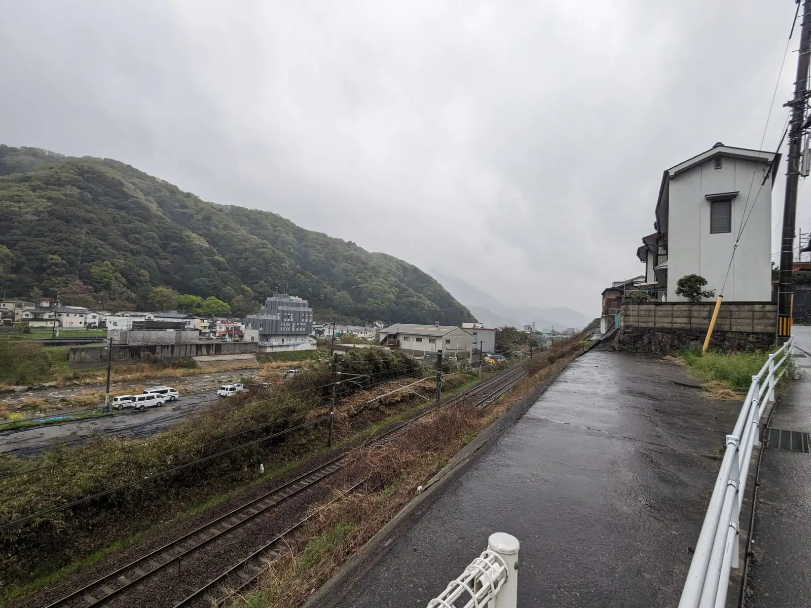 A wet road running beside a railway line and houses, with misty hills beyond