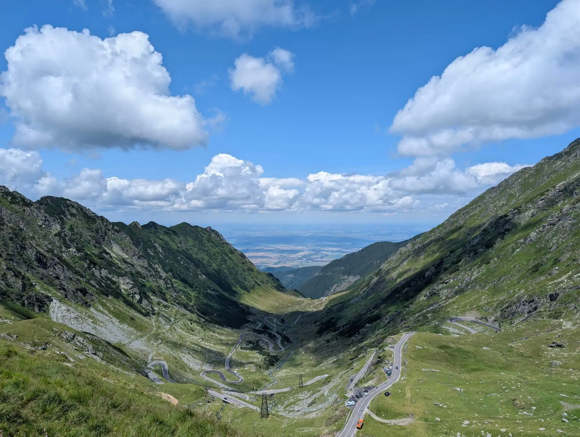 A winding road descending into a wide green mountain valley under blue sky and clouds
